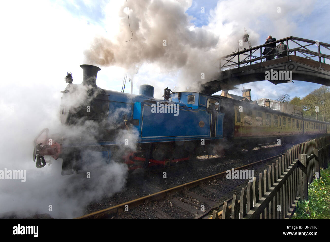 A blue steam locomotive train puffs under a footbridge into Haverthwaite station in the Lake District, Cumbria England UK Stock Photo