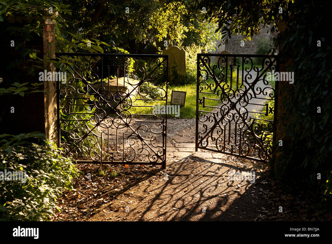 Evening sunlight casting its pattern shadow of the partly open wrought iron gate across the path leading to Bosham Churchyard. Stock Photo