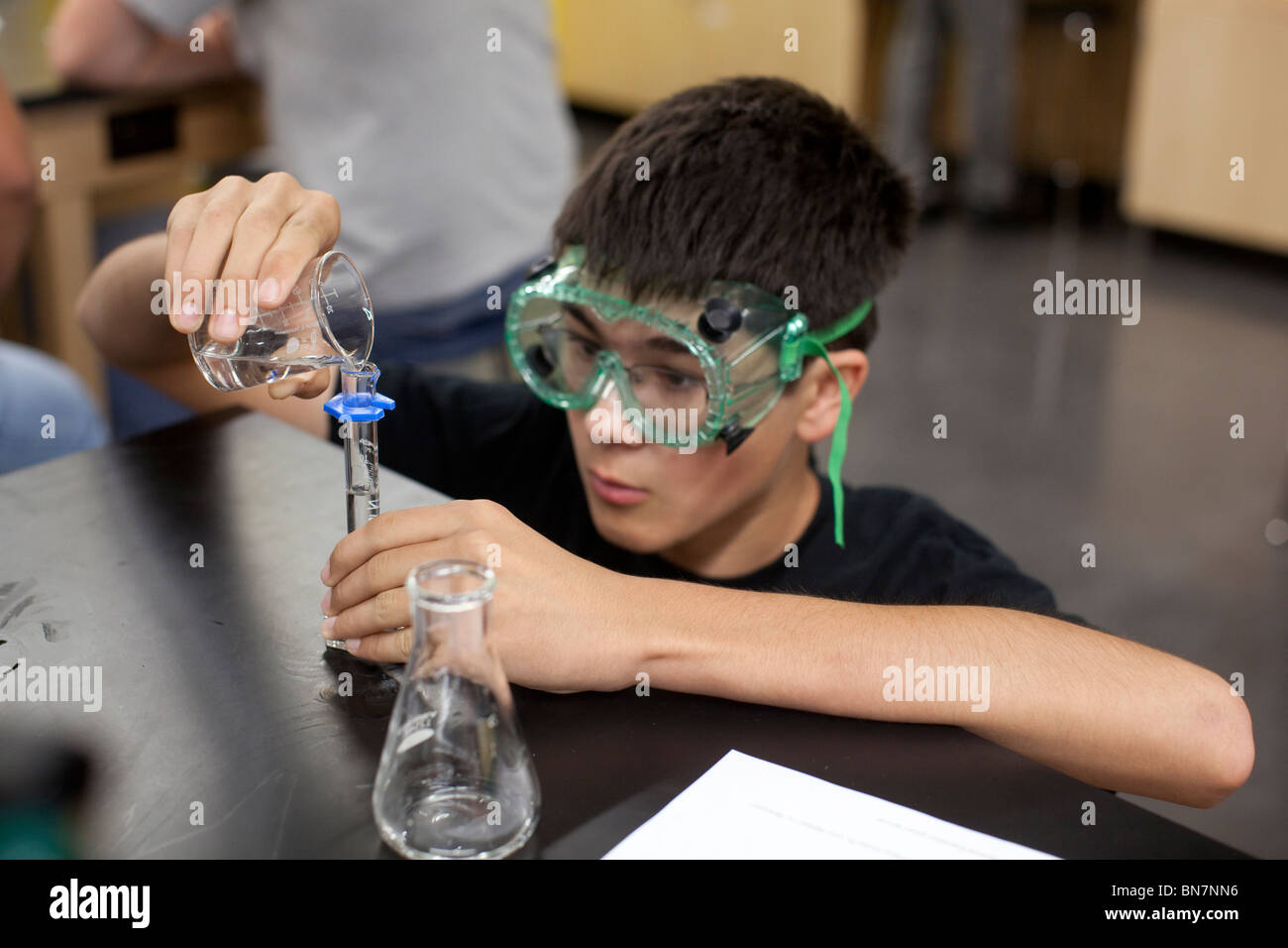Anglo male student wearing safety goggles pours solution from beaker