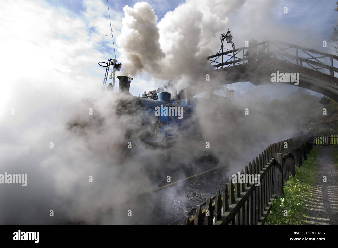 A steam locomotive train puffs under a footbridge at Haverthwaite station in the Lake District, Cumbria England UK Stock Photo