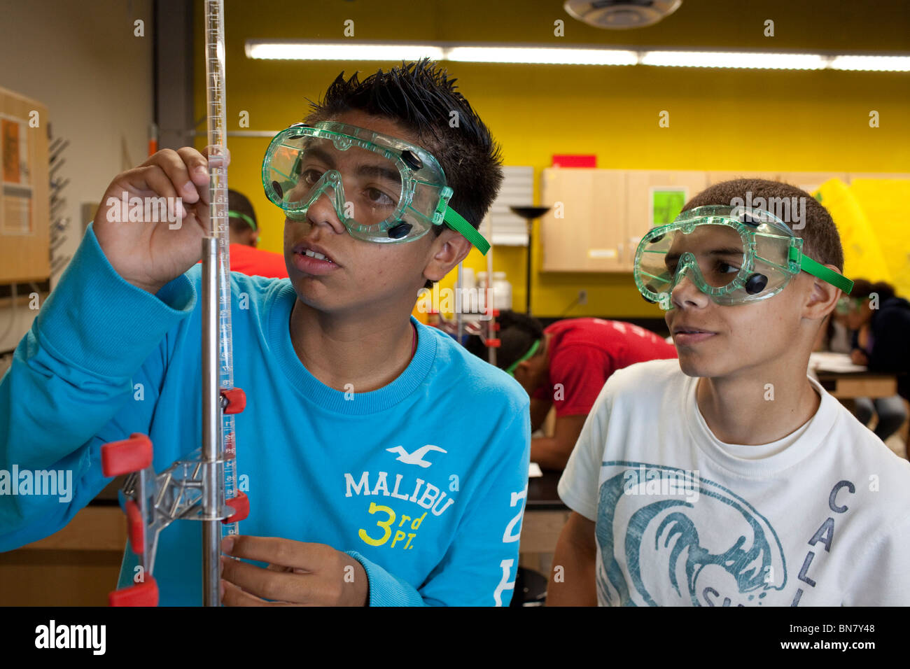 Hispanic high school boys wearing safety goggles measure solution in a graduated cylinder during titration in chemistry class. Stock Photo