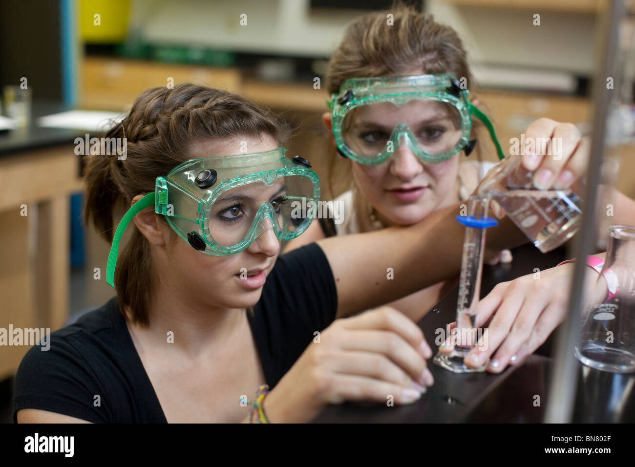 Female high school students wearing safety goggles pour liquid from a beaker into a graduated cylinder during chemistry class Stock Photo