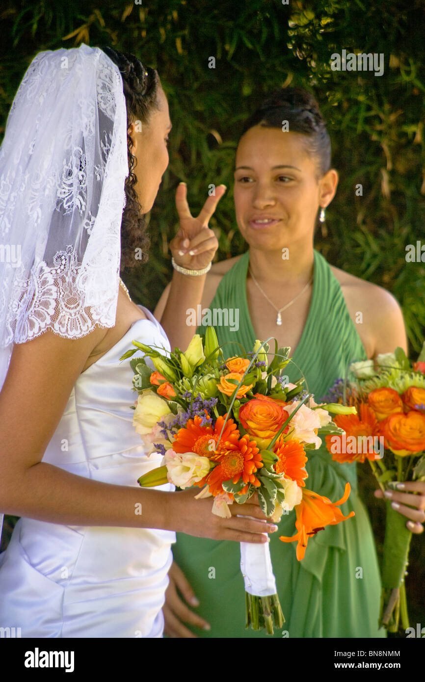 mixed-race bride makes a 'V for Victory' gesture to her bridesmaid before a formal outdoor wedding in Orange, CA. MODEL RELEASE Stock Photo