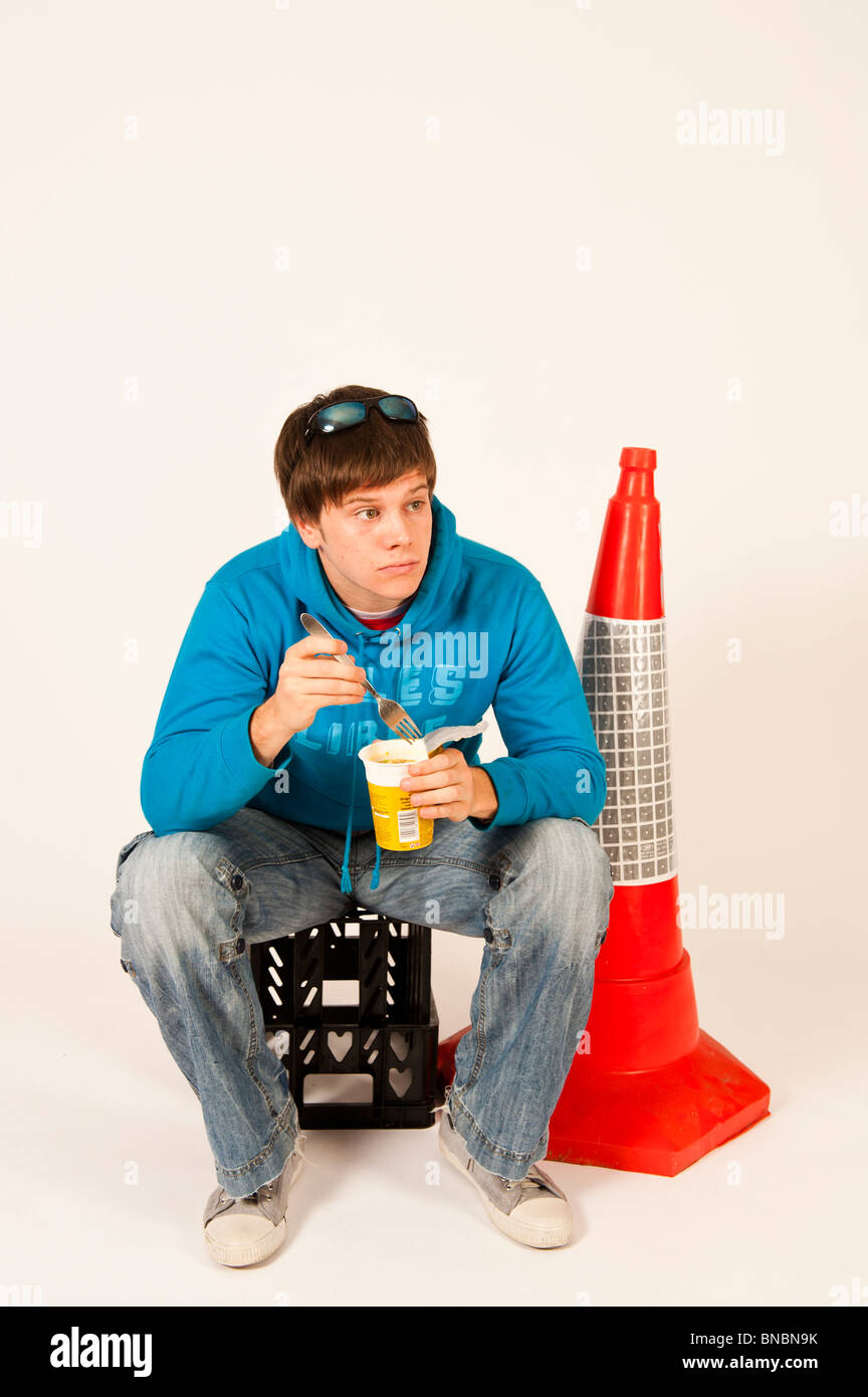 A young man eating pot noodle hot snack sitting on a plastic crate next to an orange traffic cone, UK Stock Photo