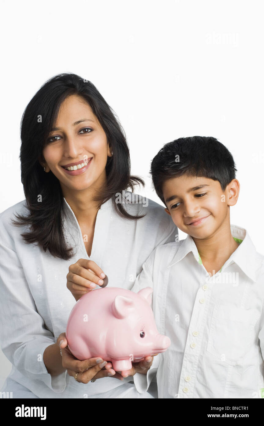 Boy inserting a coin into a piggy bank held by his mother Stock Photo ...