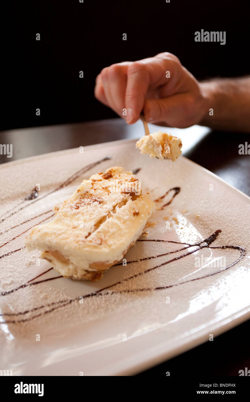 A person enjoying a slice of creamy Napoleon cake, garnished with powdered sugar and chocolate syrup, on a white ceramic plate. Stock Photo