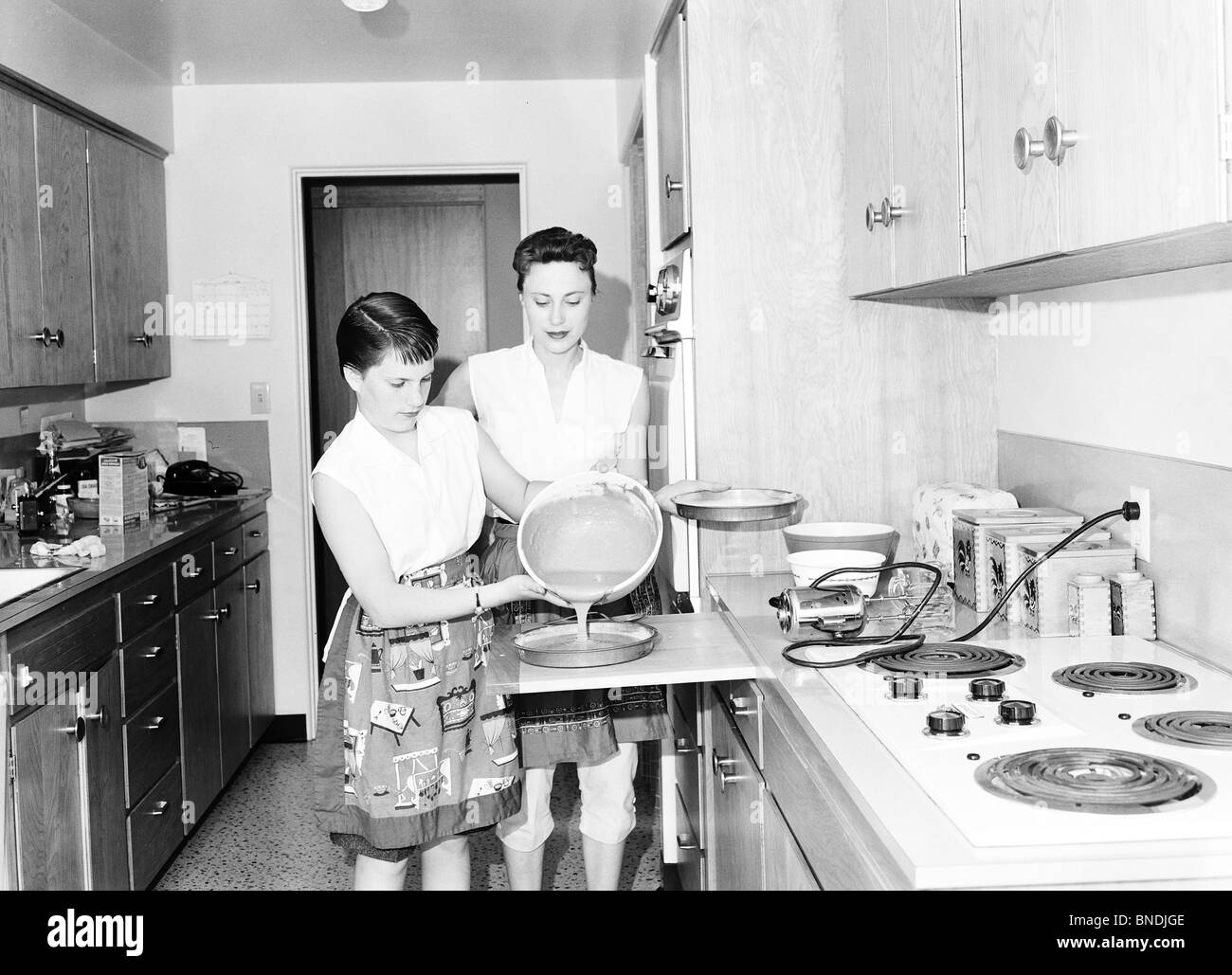 Teenage girl preparing food in the kitchen with her mother standing beside her Stock Photo