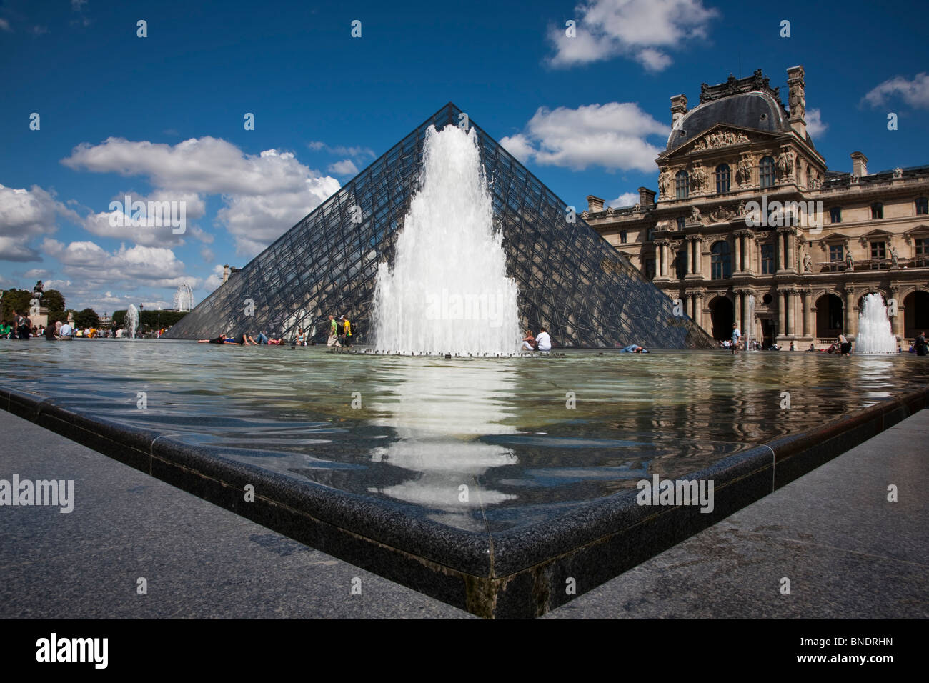 Low wide angle perspective of Fountain, Glass Pyramid, buildings at world famous Louvre museum in Paris France under blue sky and puffy clouds Stock Photo