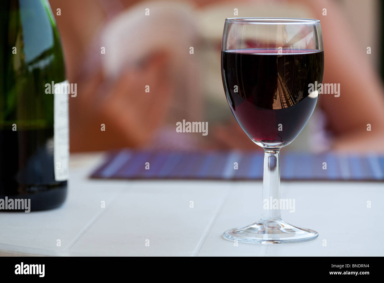 A glass of red wine on an outside table. A woman can be seen relaxing with a book in the background, Cannes france Stock Photo