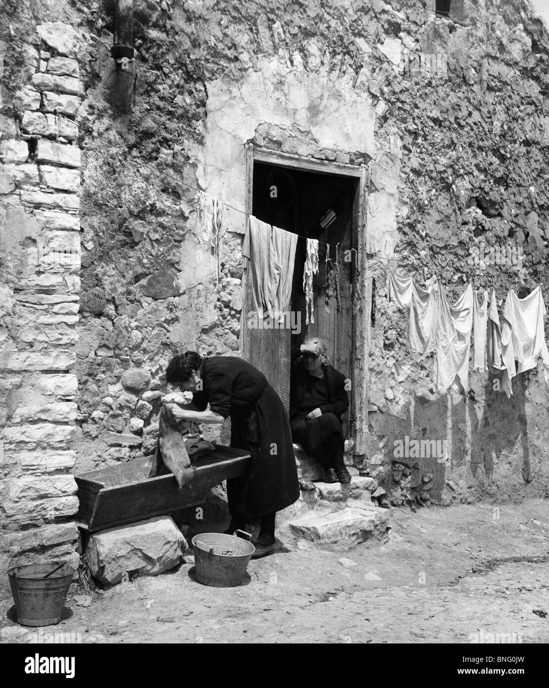 Italy, Sicily, woman doing laundry, another woman sitting near entrance Stock Photo