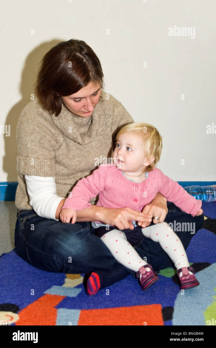 Vertical portrait of a young mum with her baby daughter at a Parent and Toddler 'sing and sign' class, signing the word Daddy. Stock Photo