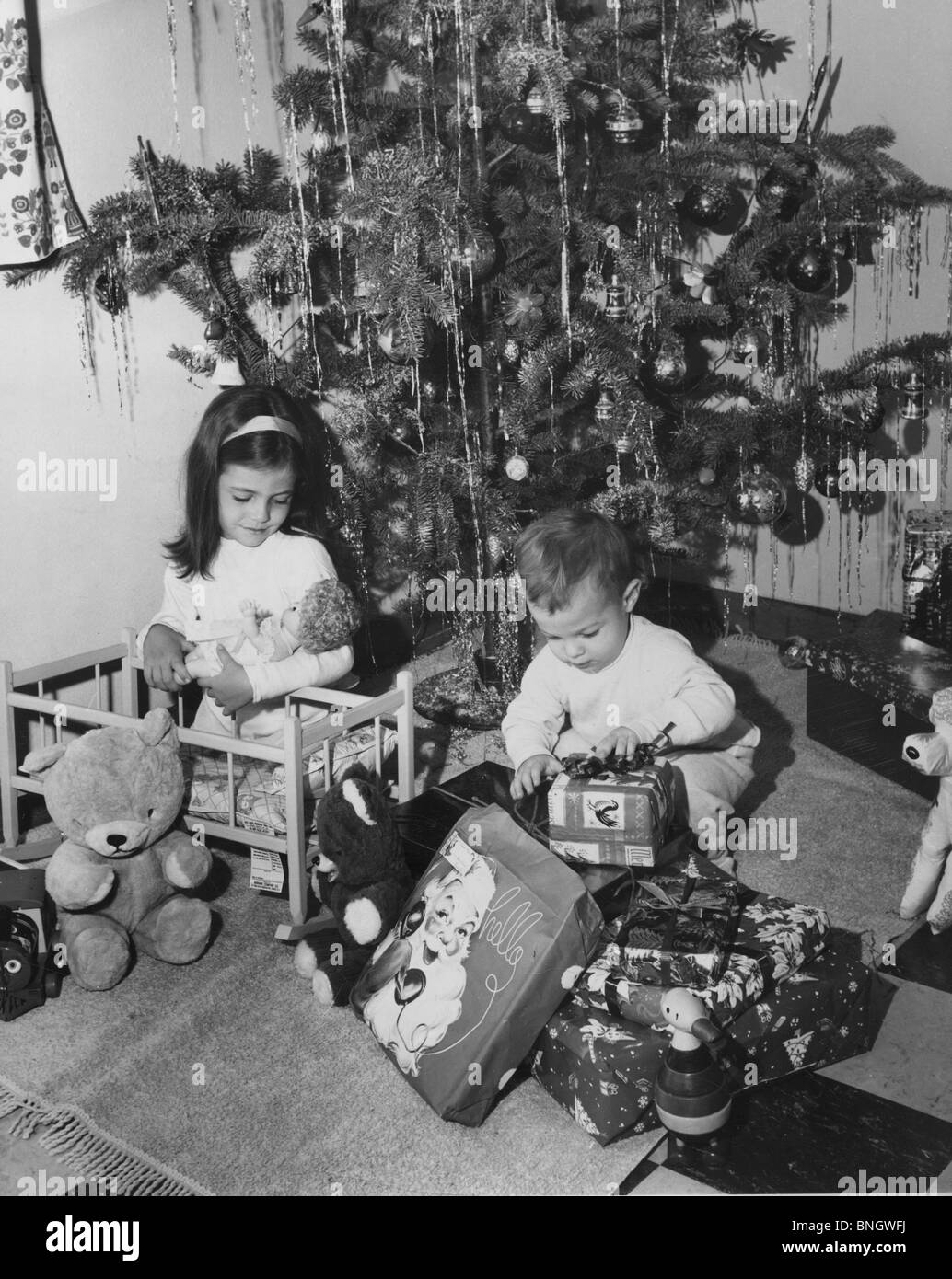 Girl playing with a doll and her brother opening a Christmas present beside her, 1960s Stock Photo