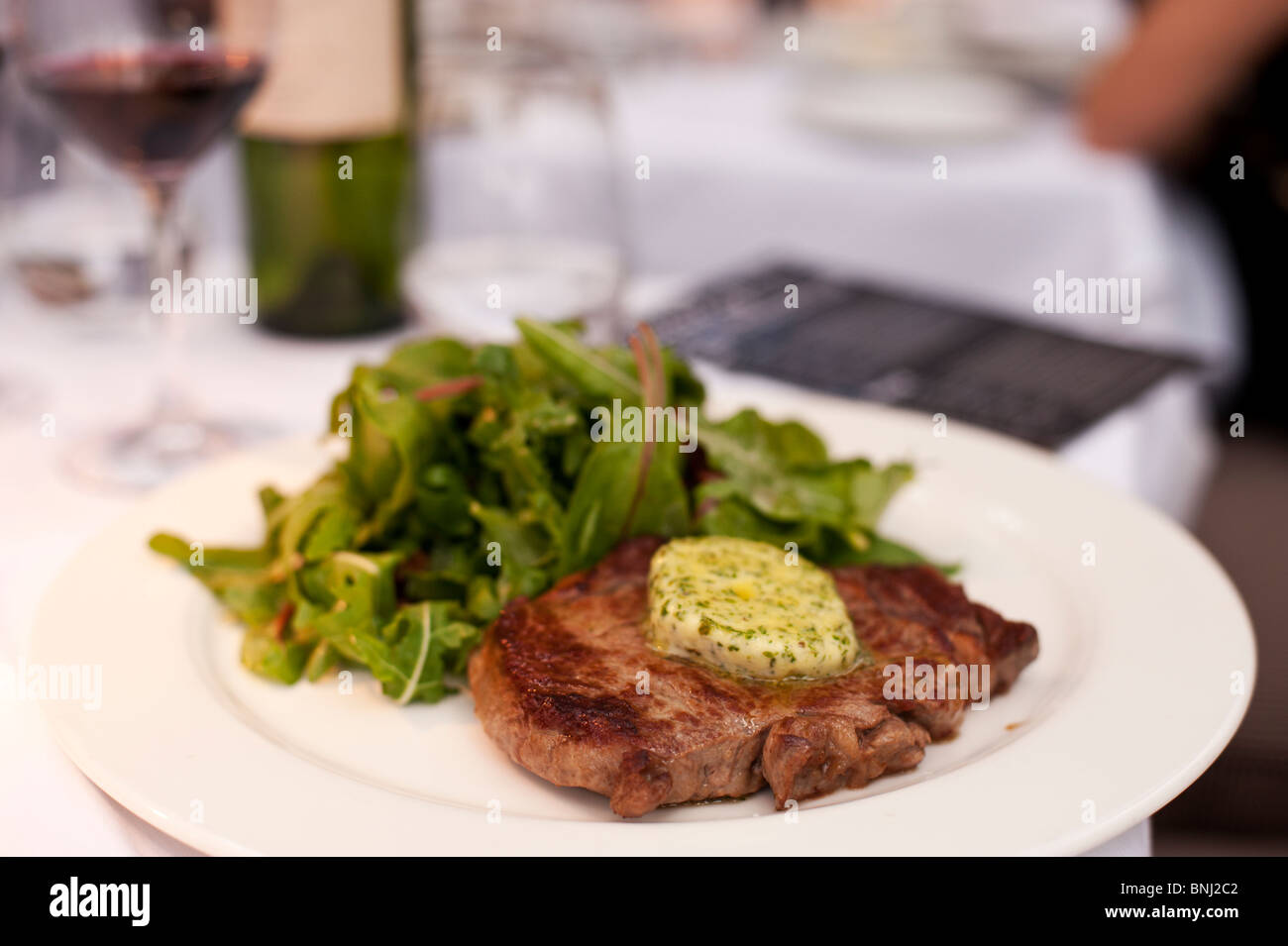 Steak served with herb butter and a green salad, with red wine in the background, on the white table linen at Gee's Restaurant. Stock Photo