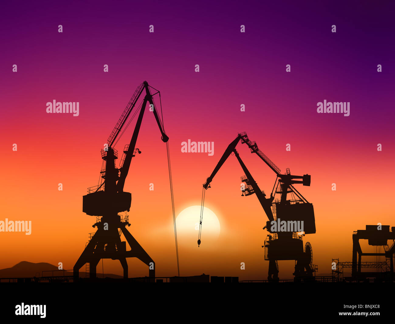 Two cranes sit dramatically against a colorful sunset in a large shipyard Stock Photo