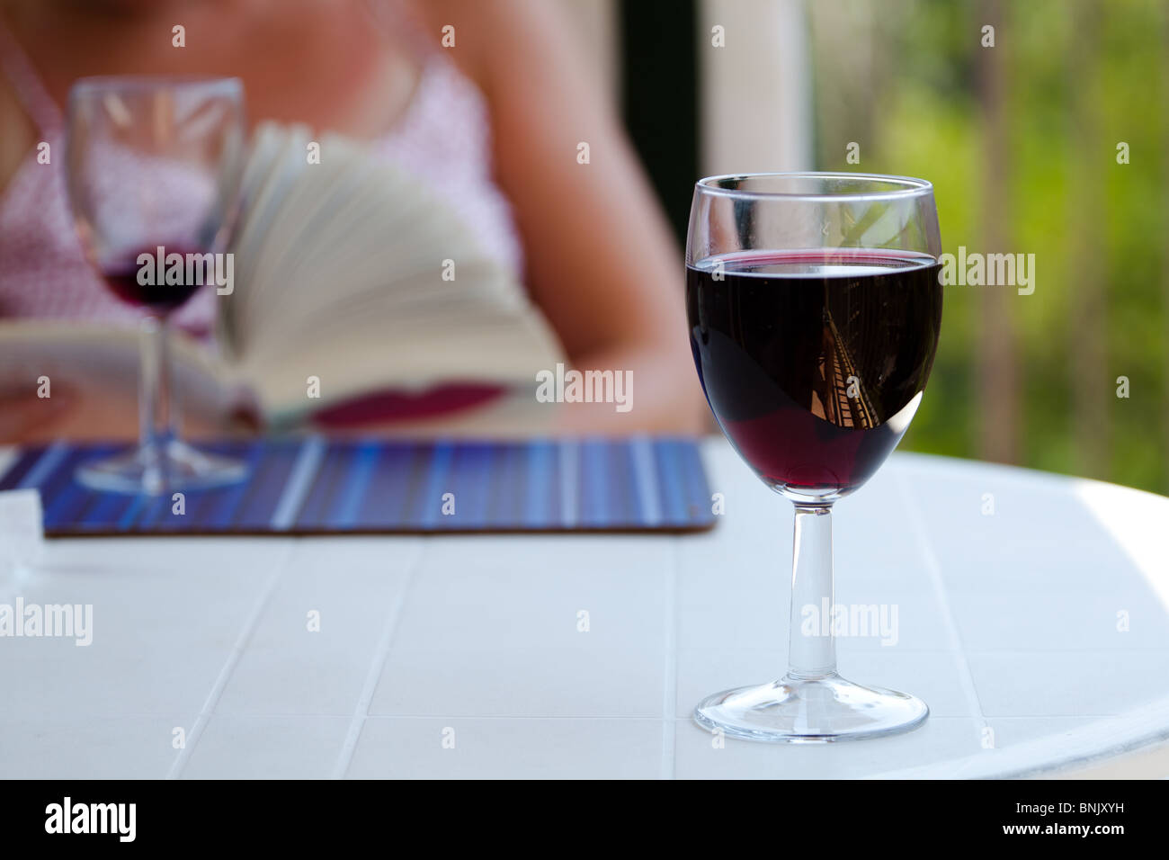 A glass of red wine on an outside table with a woman relaxing in the background reading a book Stock Photo