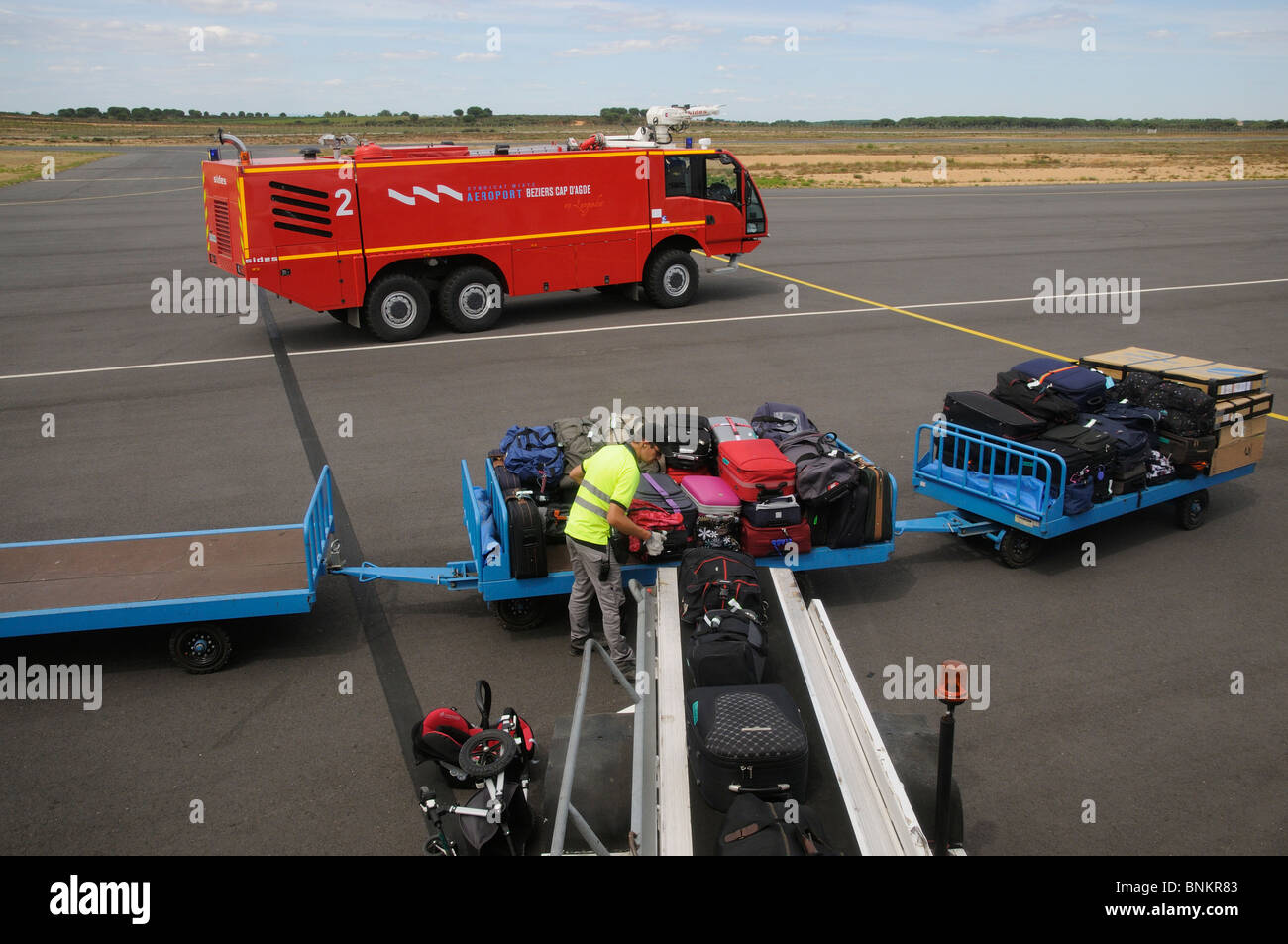 Airport services a fire truck & baggage handler loading bags from a trolley onto an aircraft at Beziers International Airport EU Stock Photo