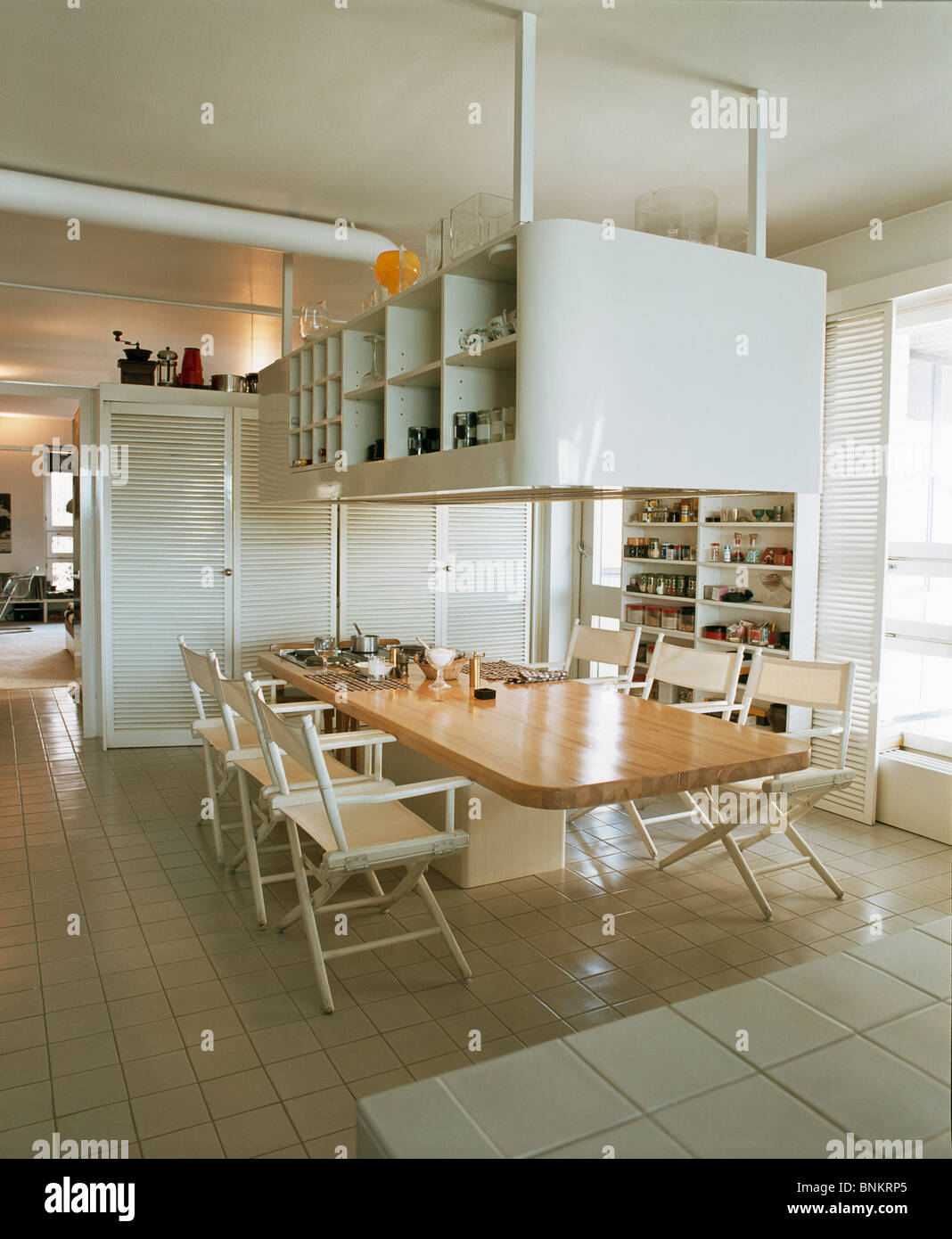 Suspended shelving above island unit with long fitted table and cream  director's chairs in modern white kitchen with tiled floor Stock Photo -  Alamy