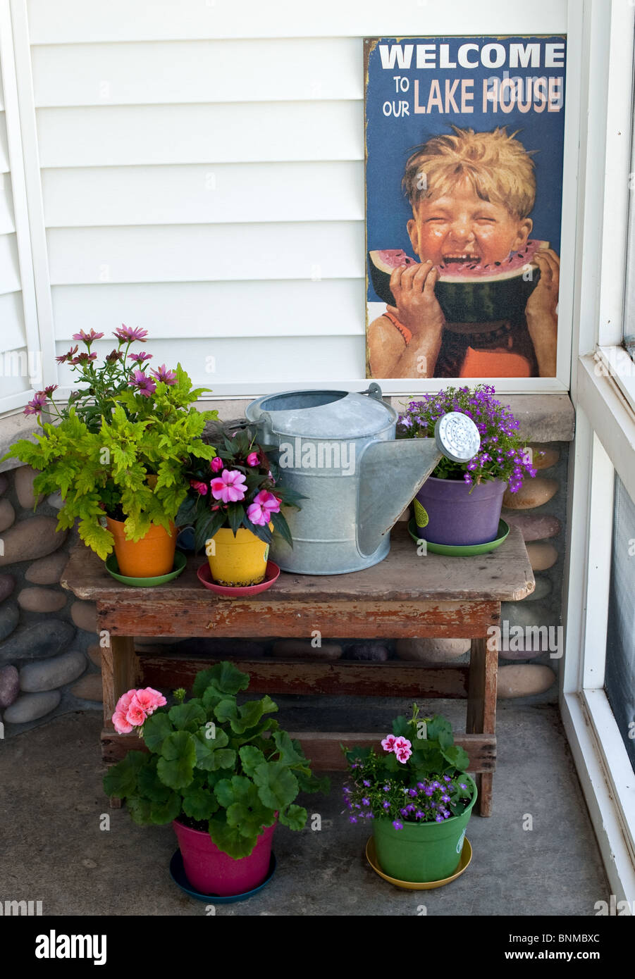 Lake porch with flower on a bench with a tin watering can and a welcoming sign. Stock Photo