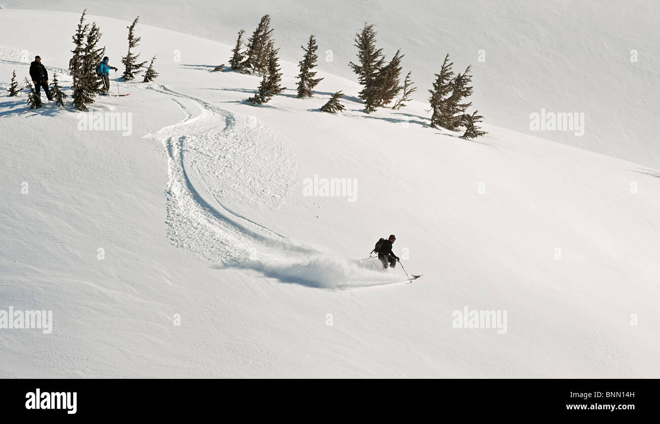 A skier and snowboarder watch as a fellow backcountry skier starts a descent in Turnagain Pass, Alaska Stock Photo