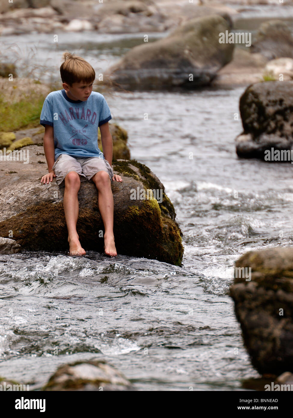 Young boy sat on a rock by the East Lyn river,  Lynmouth, Devon, UK Stock Photo