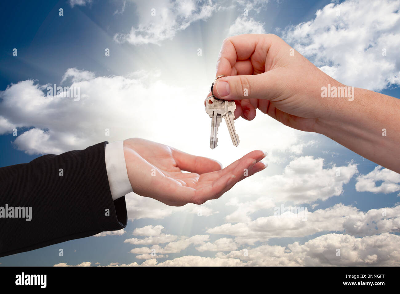 Male Hand Handing Over Keys to Female Hand Over Dramatic Clouds and Sun Rays. Stock Photo