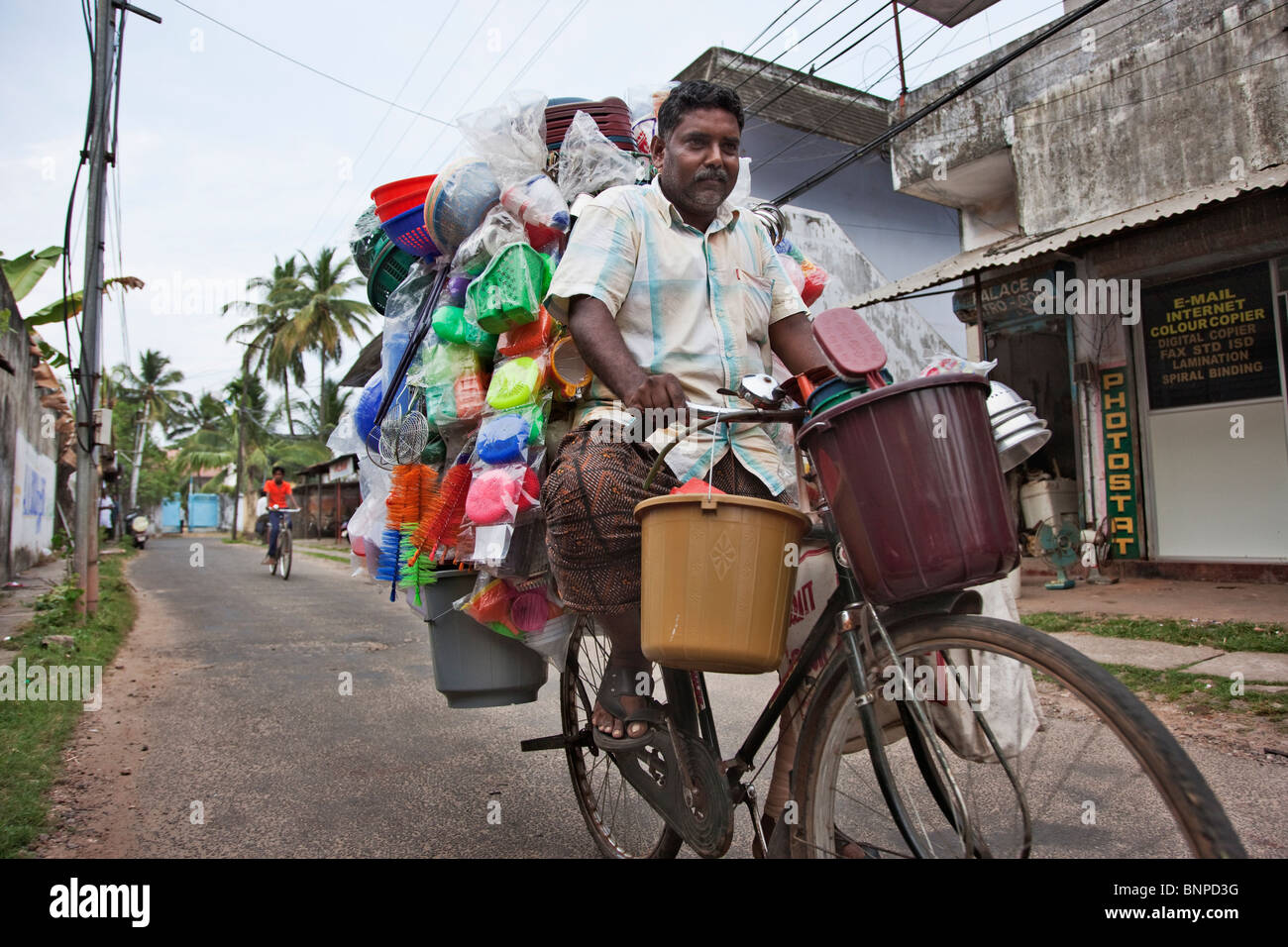 Middle aged Man on bicycle selling various types of plastic household items cycling down a rural city street Stock Photo