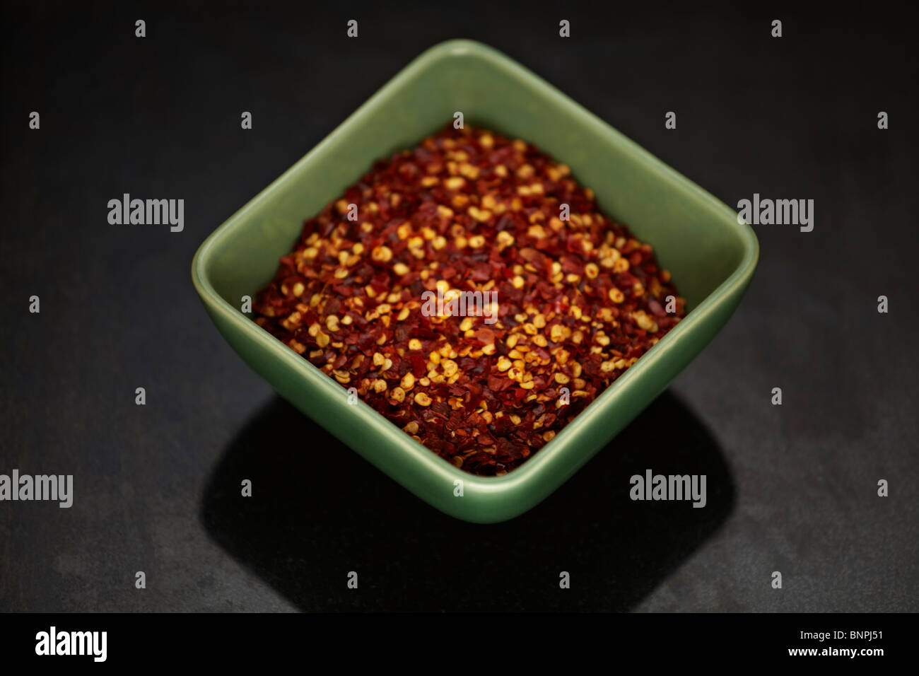 Dried chilli flakes and seeds in a green bowl on a black marble surface Stock Photo