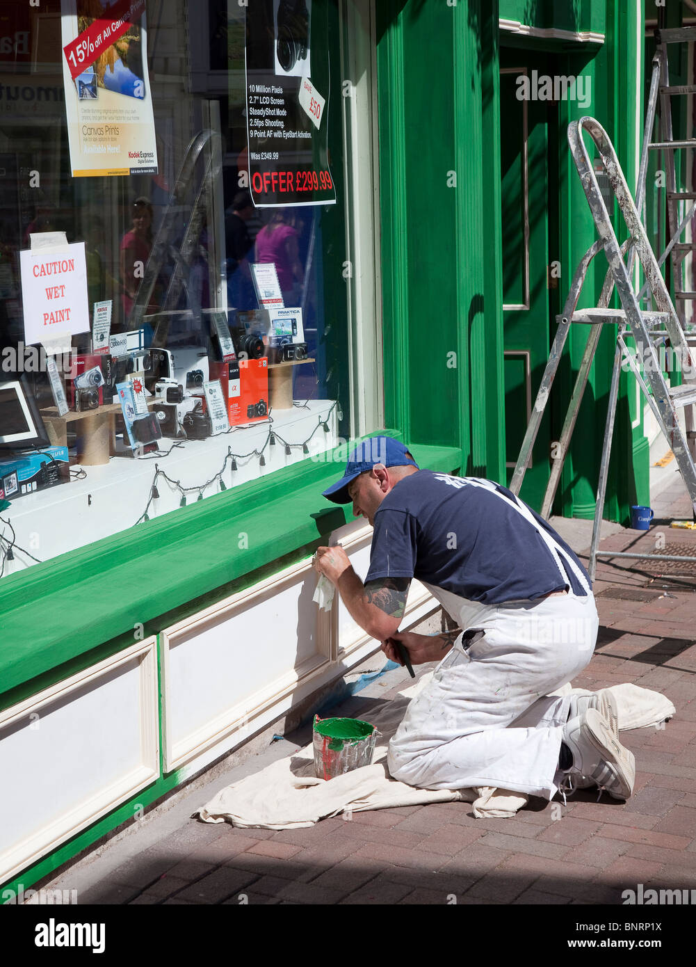 Man painting shop windowframe with caution wet paint notice in window Abergavenny Wales UK Stock Photo