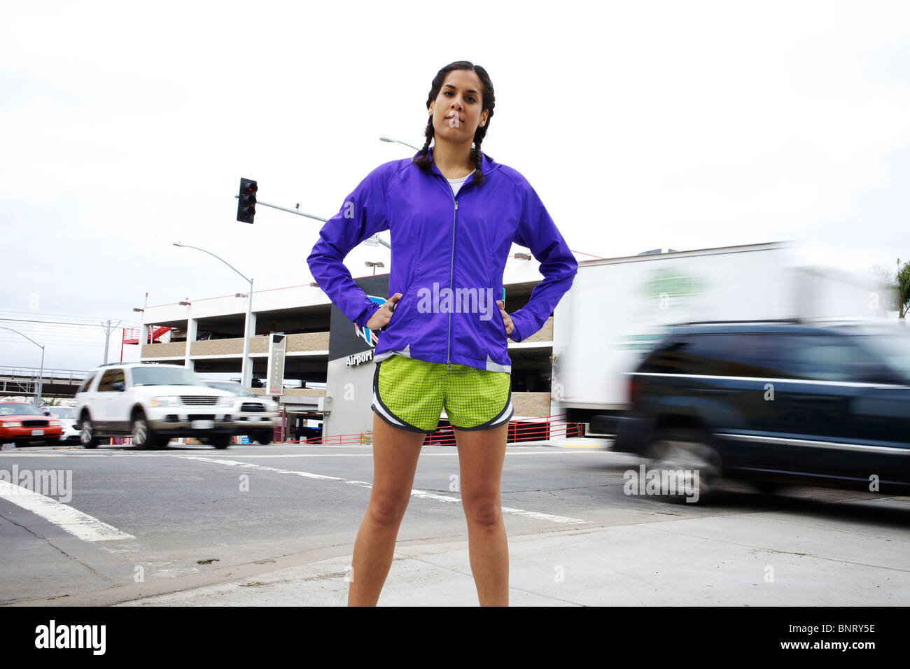 A female runner stands on the corner of two streets and cars pass by in