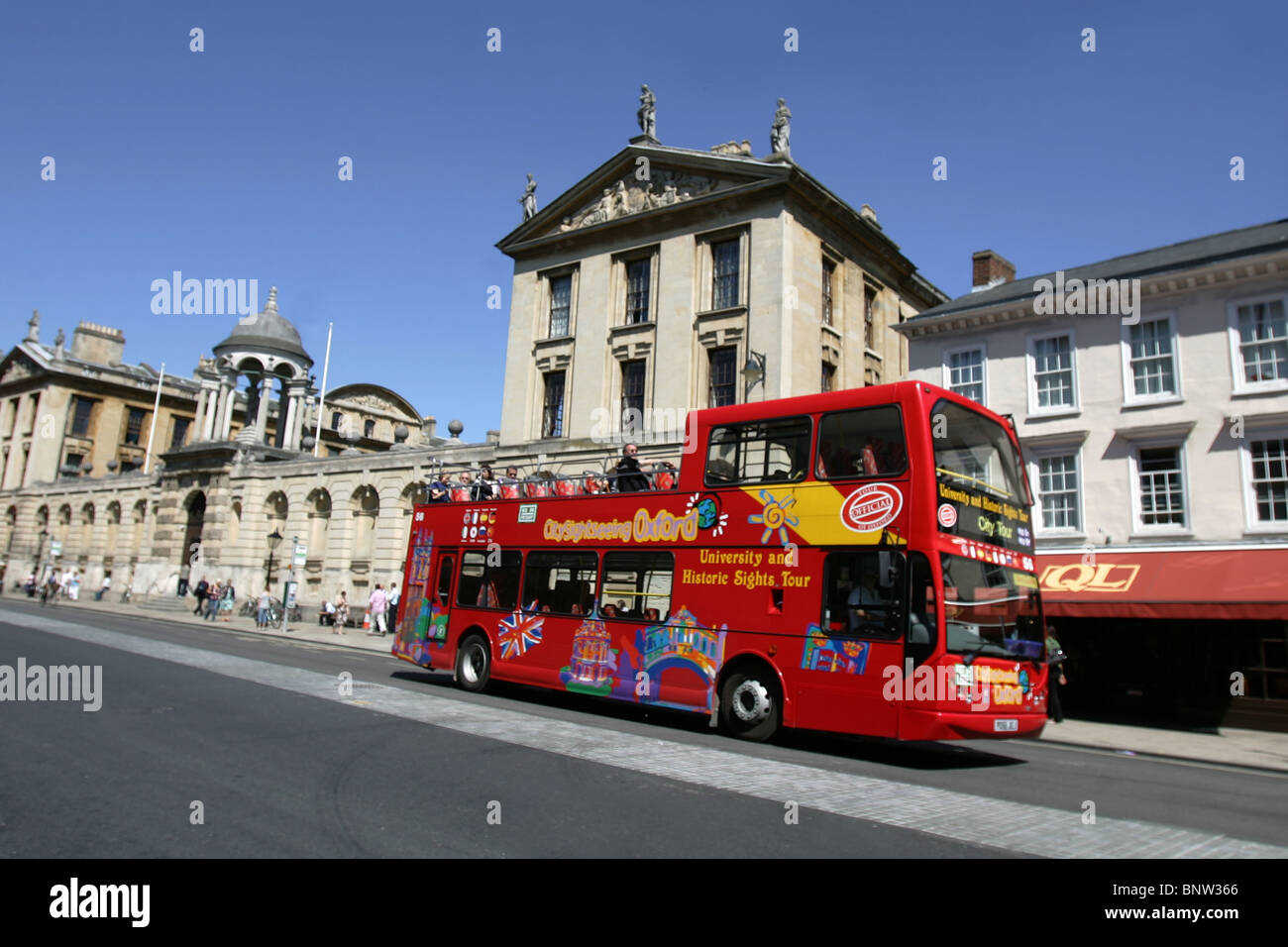Red sightseeing tour bus drives past The Queen's College, Oxford City. Stock Photo