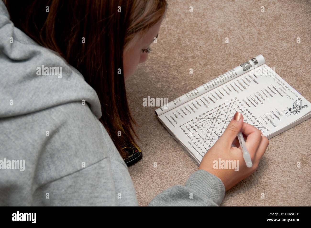 A teenage brown haired Caucasian girl lies on the carpet and works a word puzzle. USA. Stock Photo