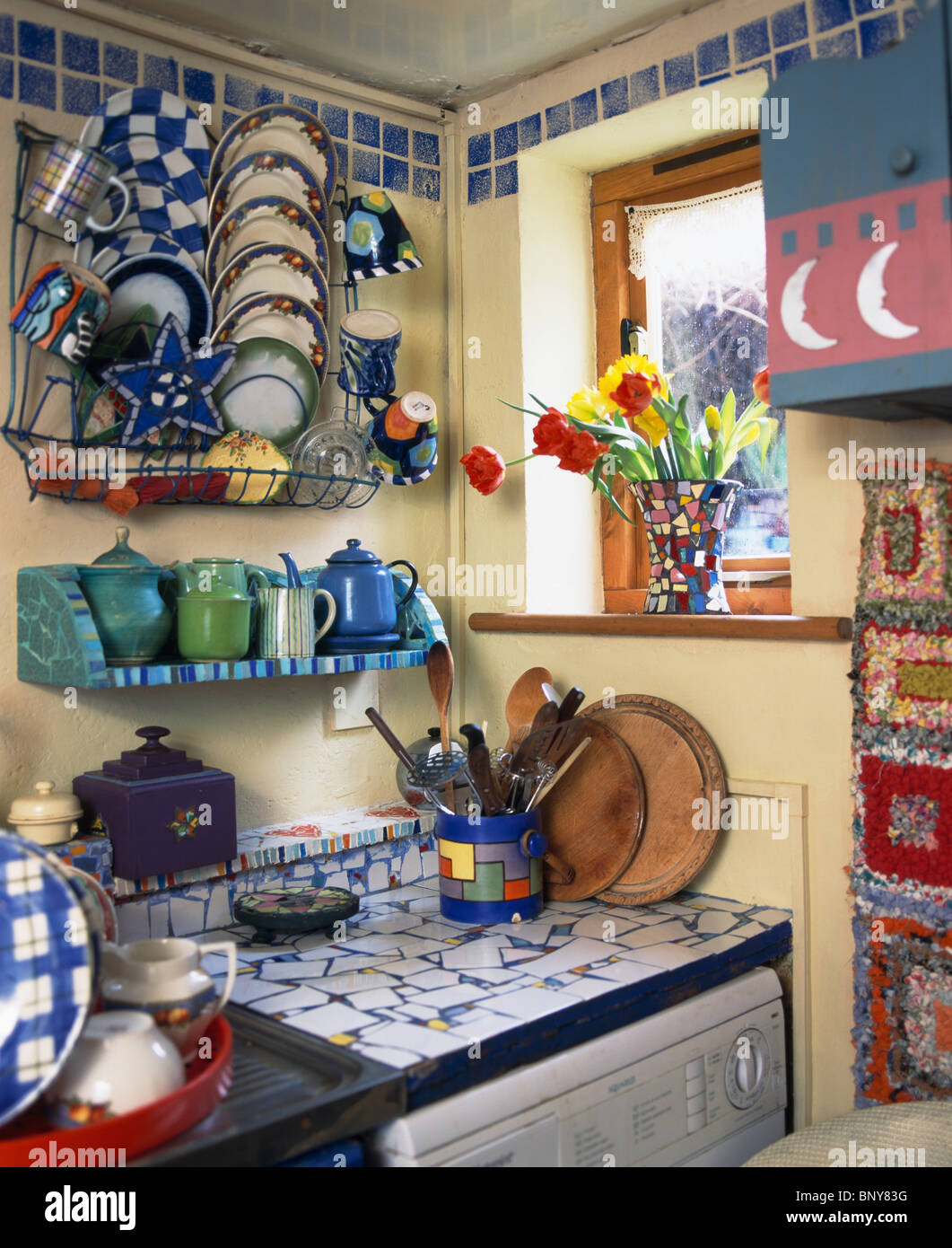 Close-up of colorful china and metal pots on metal racks above mosaic worktop in small cluttered cottage kitchen Stock Photo
