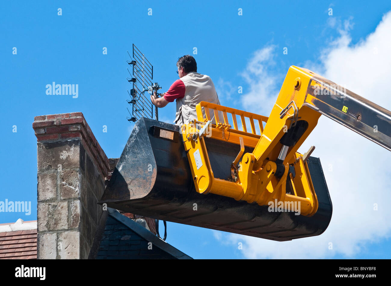 Television aerial being fitted to chimney by man standing (no safety equipment) in JCB Telescopic Handler bucket - France. Stock Photo