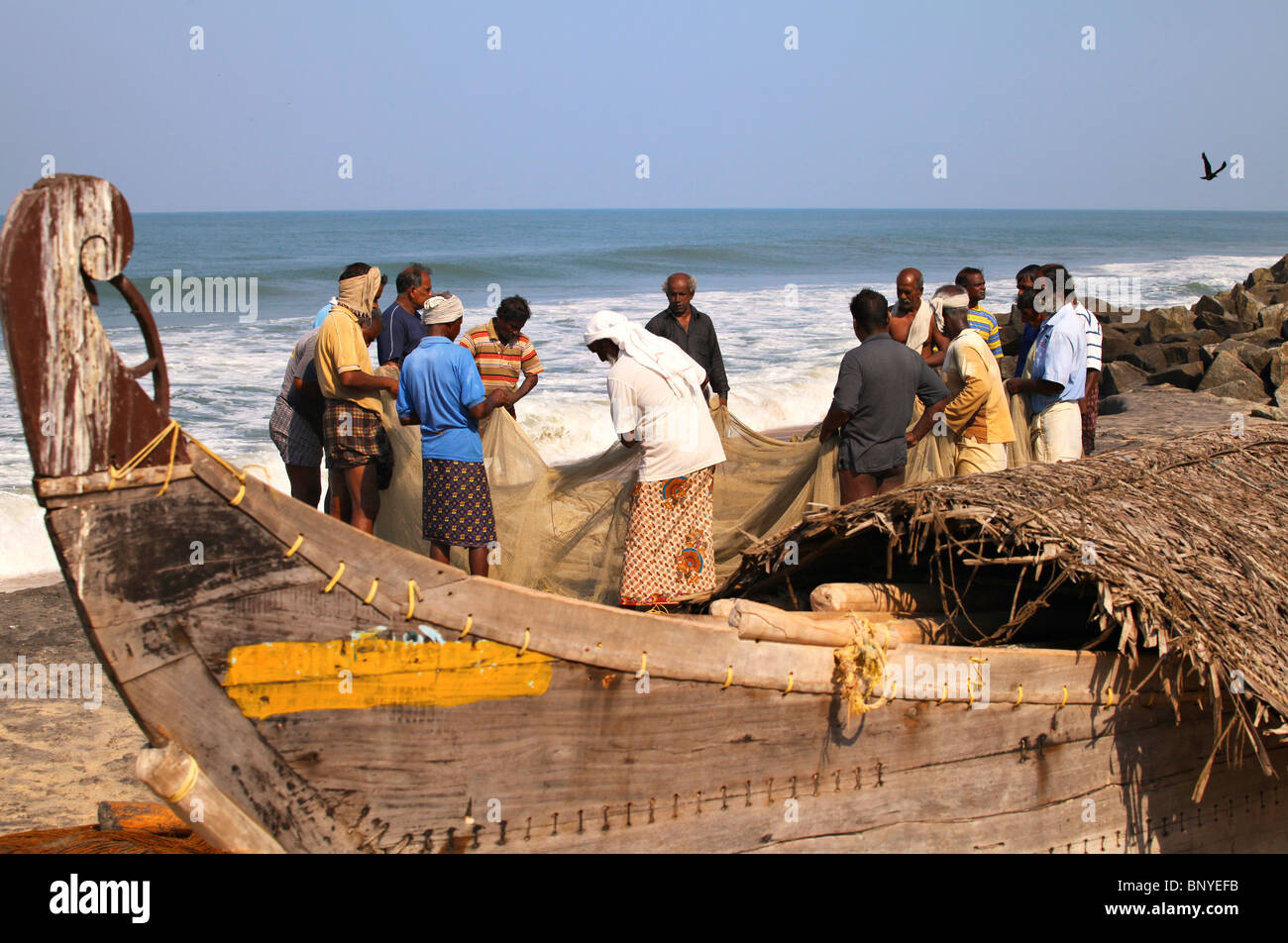 Indian fisherman work on their nets next to a traditional Keral fishing boat, with its planks joined by stitching. Stock Photo