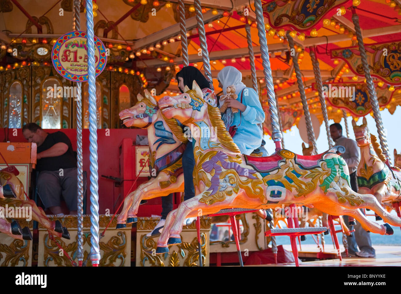 Girls in muslim headscarfs, one holding a teddy bear, on a fairground carousel, on Brighton's seafront Stock Photo