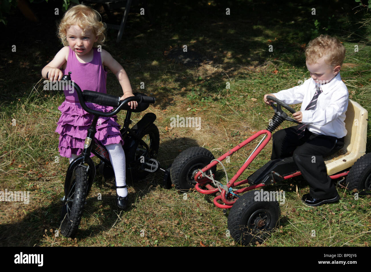 Little girl boy siblings toddlers playing together in party clothes dressed up bicycle and soapbox car kart MODEL RELEASED Stock Photo