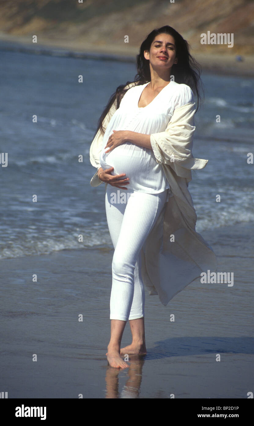 mixed race heavily pregnant woman standing on the beach smiling into