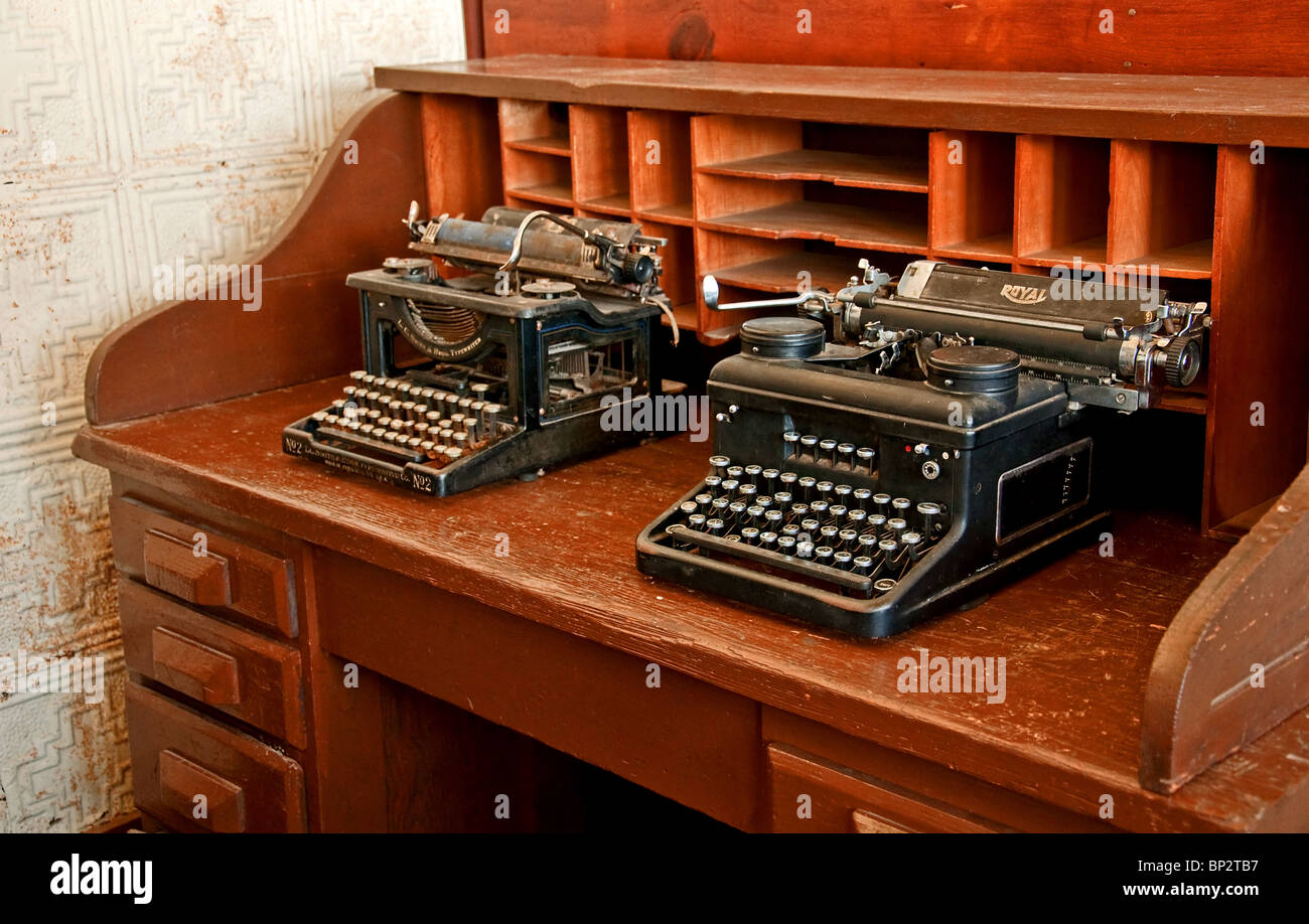 Two antique black typewriters are sitting on a vintage turn of the century wooden desk, along with vintage wall paper. Stock Photo