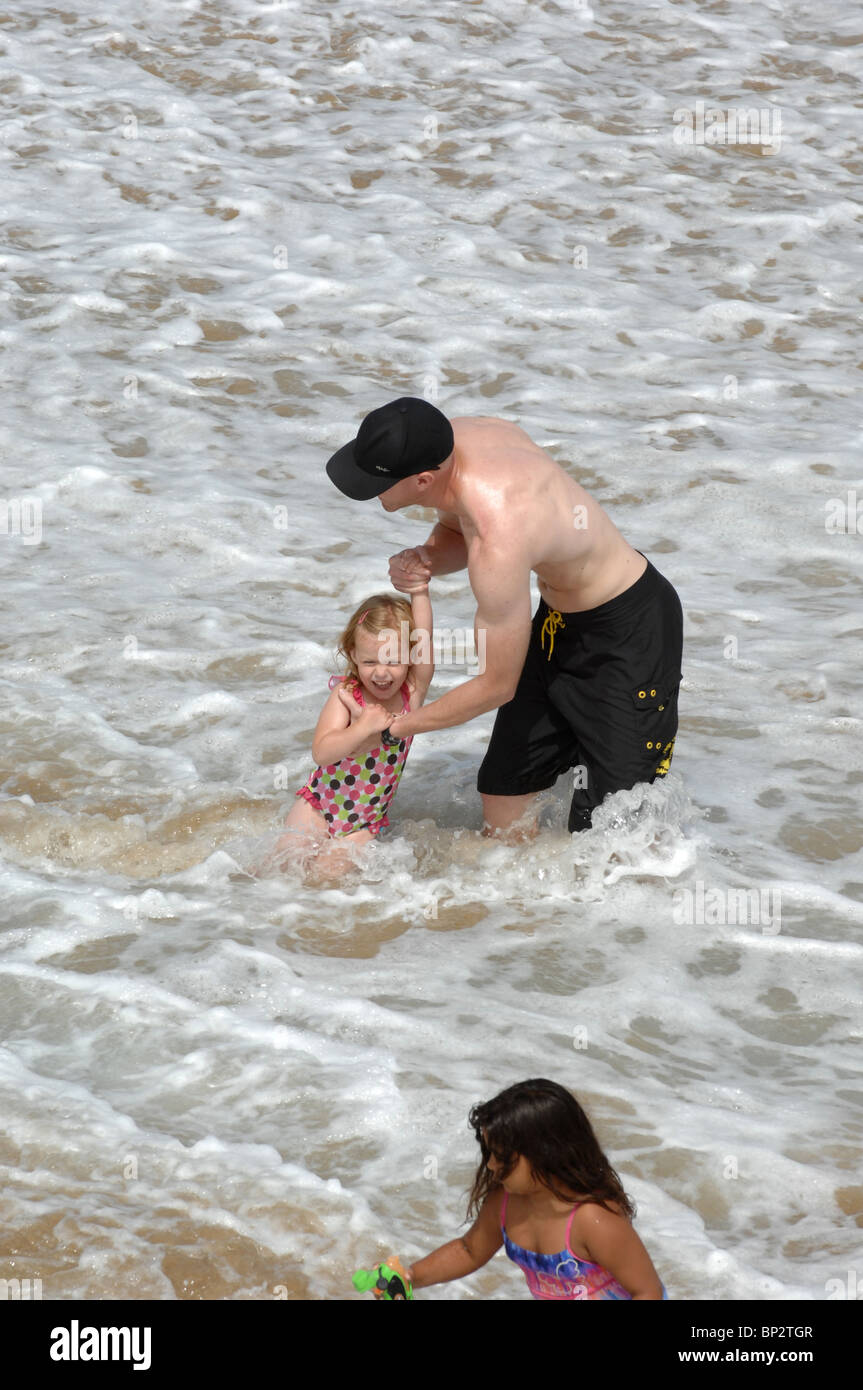 An attractive athletic man plays with his daughter in the waves at the ocean and protects her from the surf. Stock Photo