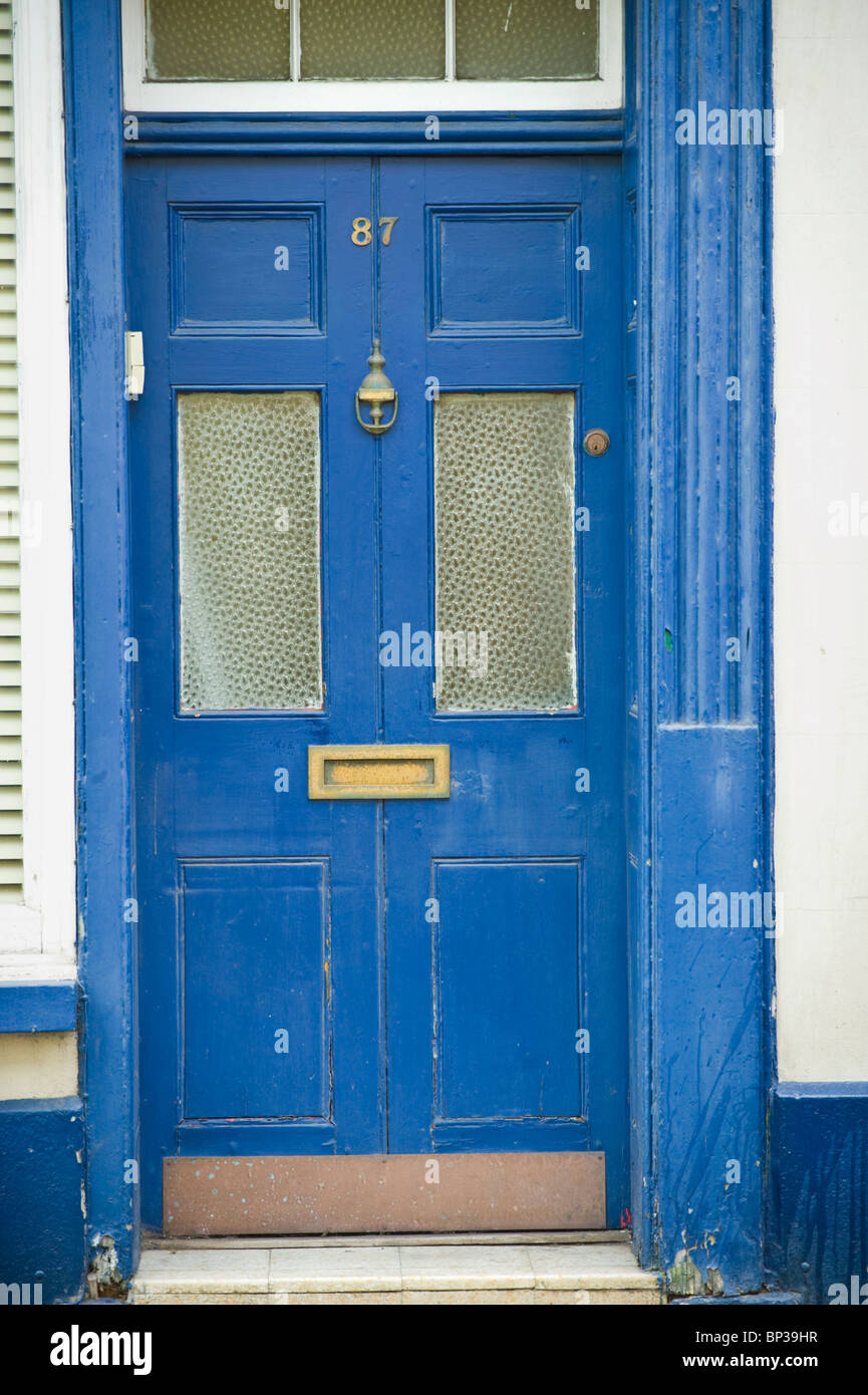 Scruffy blue painted wooden paneled glazed front door no. 87 with brass knocker letterbox and knob of house in UK Stock Photo