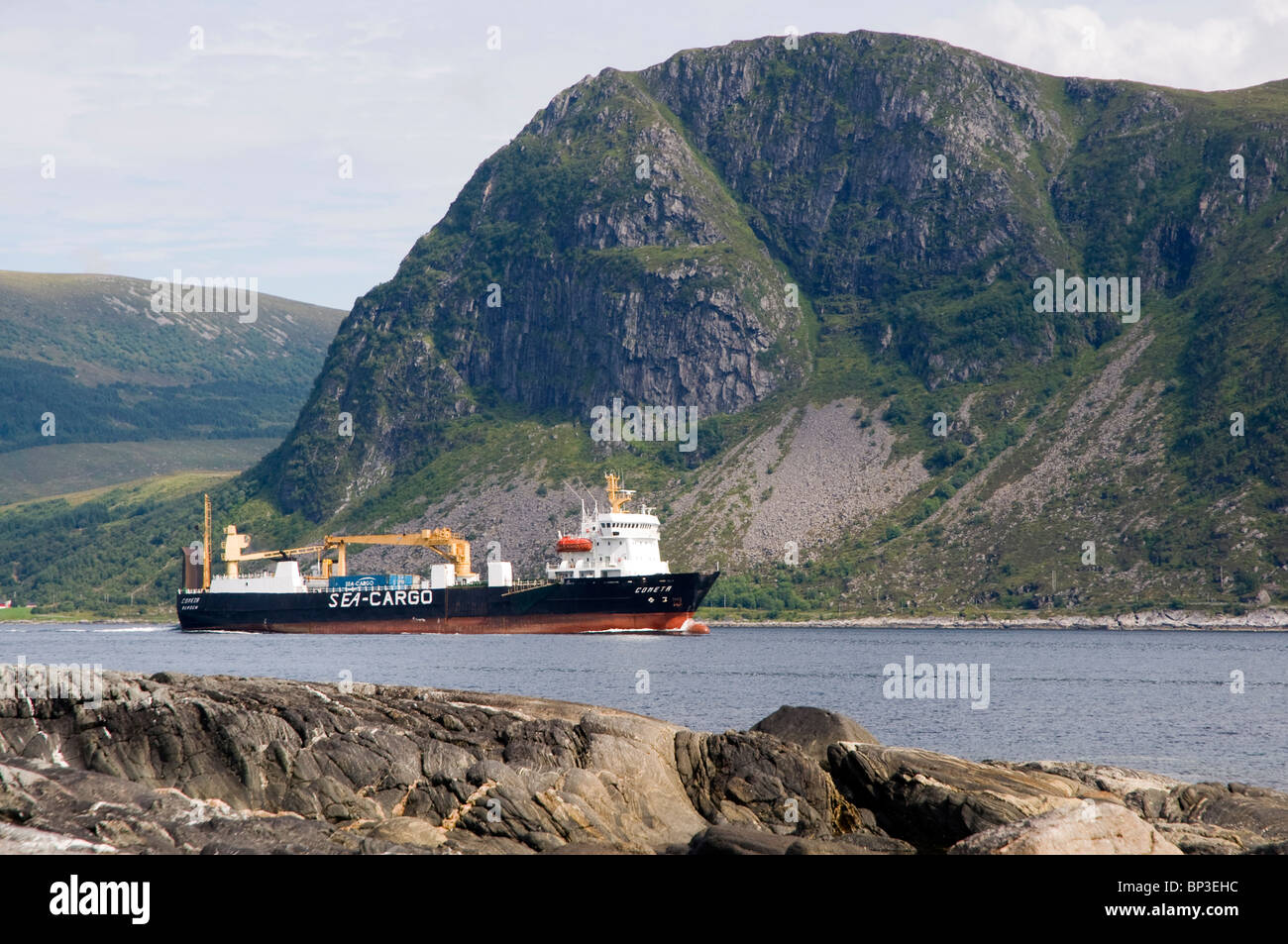 Sea cargo commercial vessel passing through the narrow sound between Sand Isle and the mainland on the way to Ulsteinvik. Stock Photo