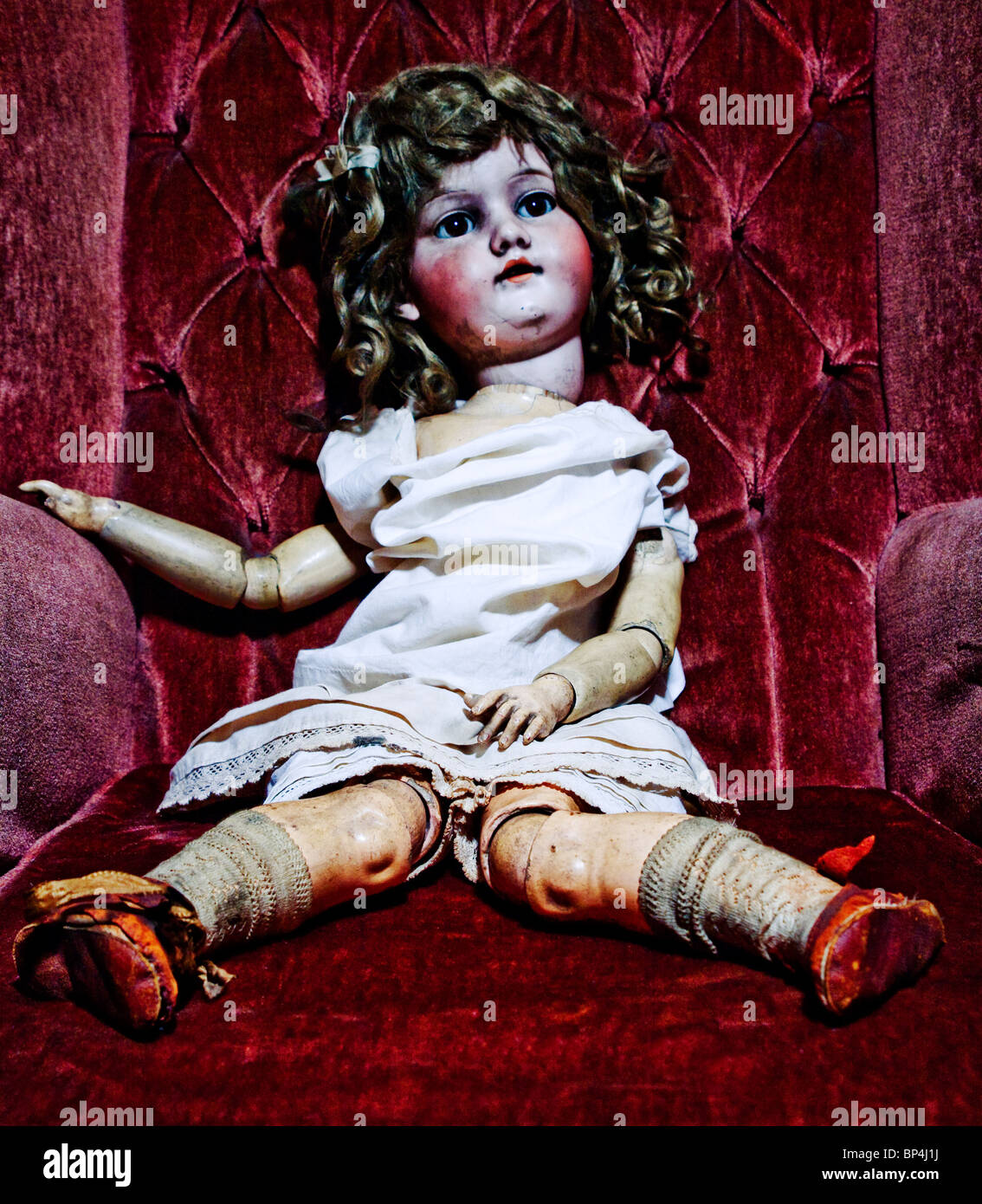 An old fashioned antique doll seated on a burgundy velvet sofa wearing a white dress. Stock Photo