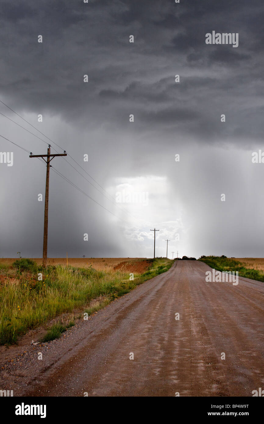Rural thunderstorm scene - dirt road leads to bright light in sky between dark  looming cumulus thunder clouds releasing rain Stock Photo