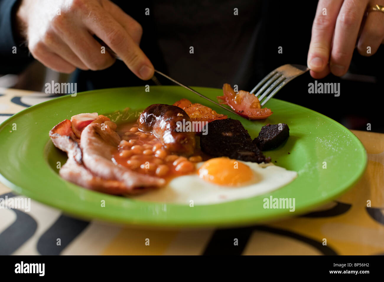 A man eats a full English breakfast (with eggs, bacon, beans, sausage, black pudding, tomato, and egg) at a cafe. Stock Photo