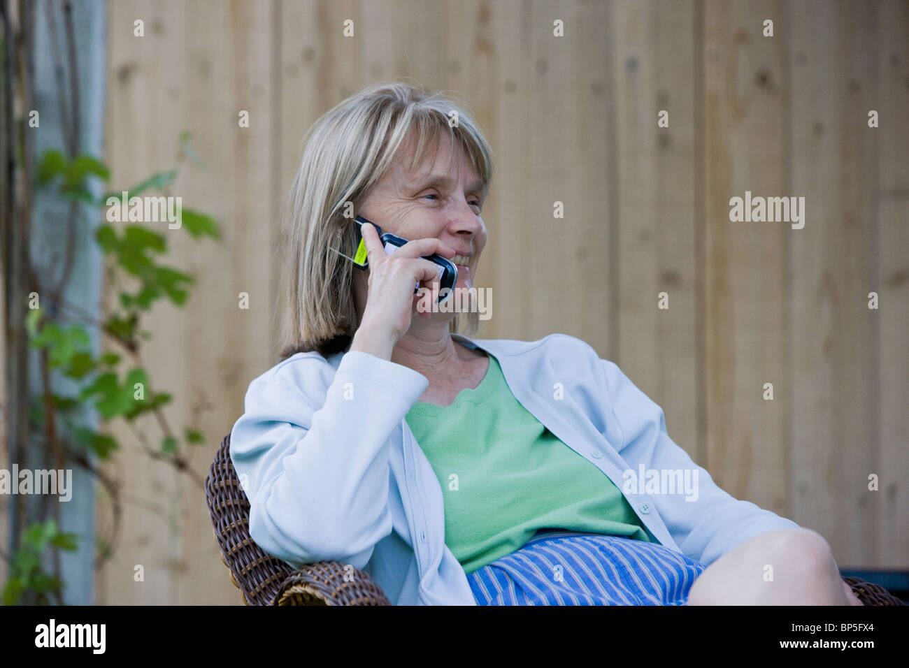 Middle aged woman talking on a telephone while seated in a wicker chair in a residential yard. Stock Photo