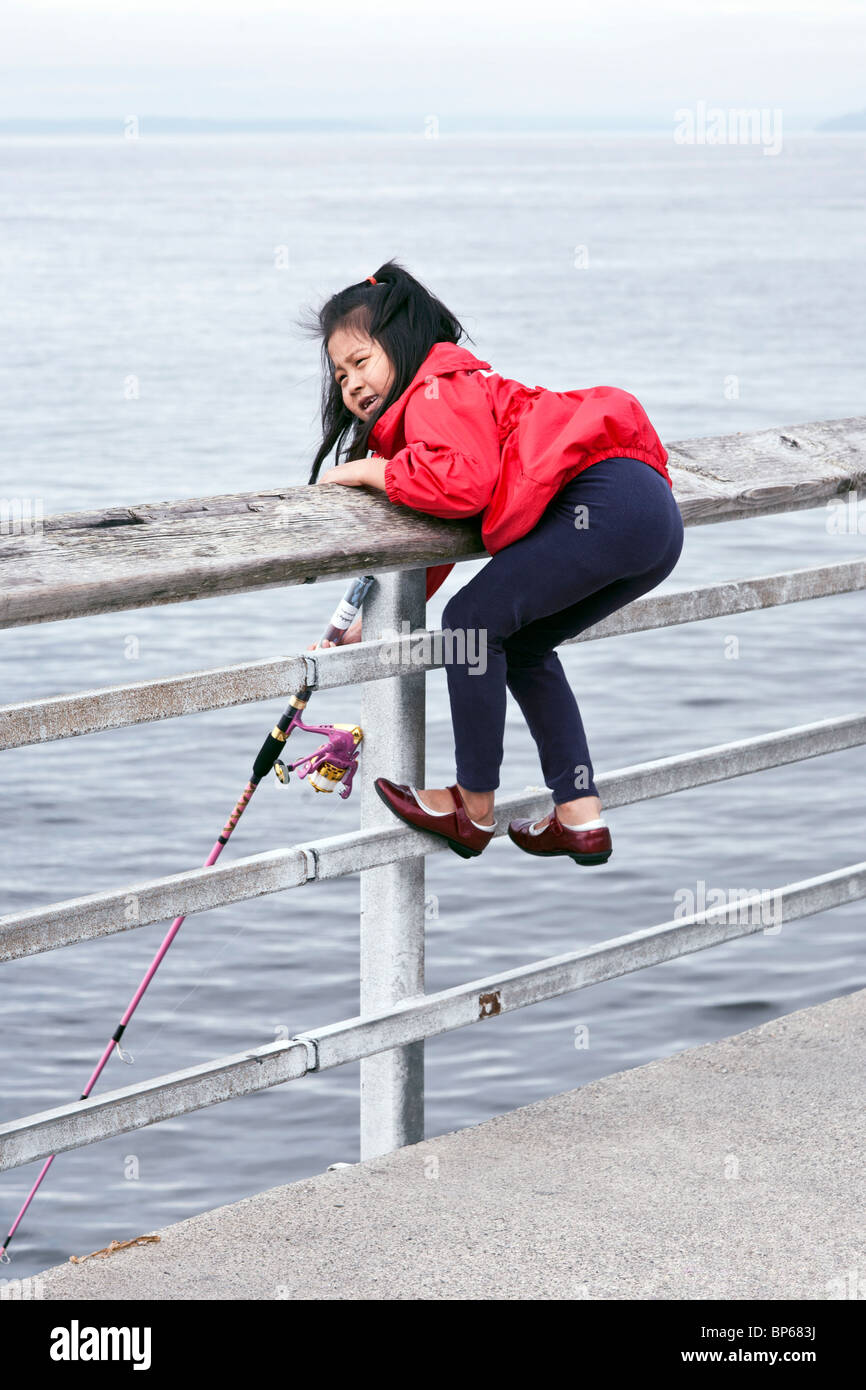 happy little Asian girl perches precariously on railing of Edmonds fishing pier with  her fanny in the air as she fishes Stock Photo