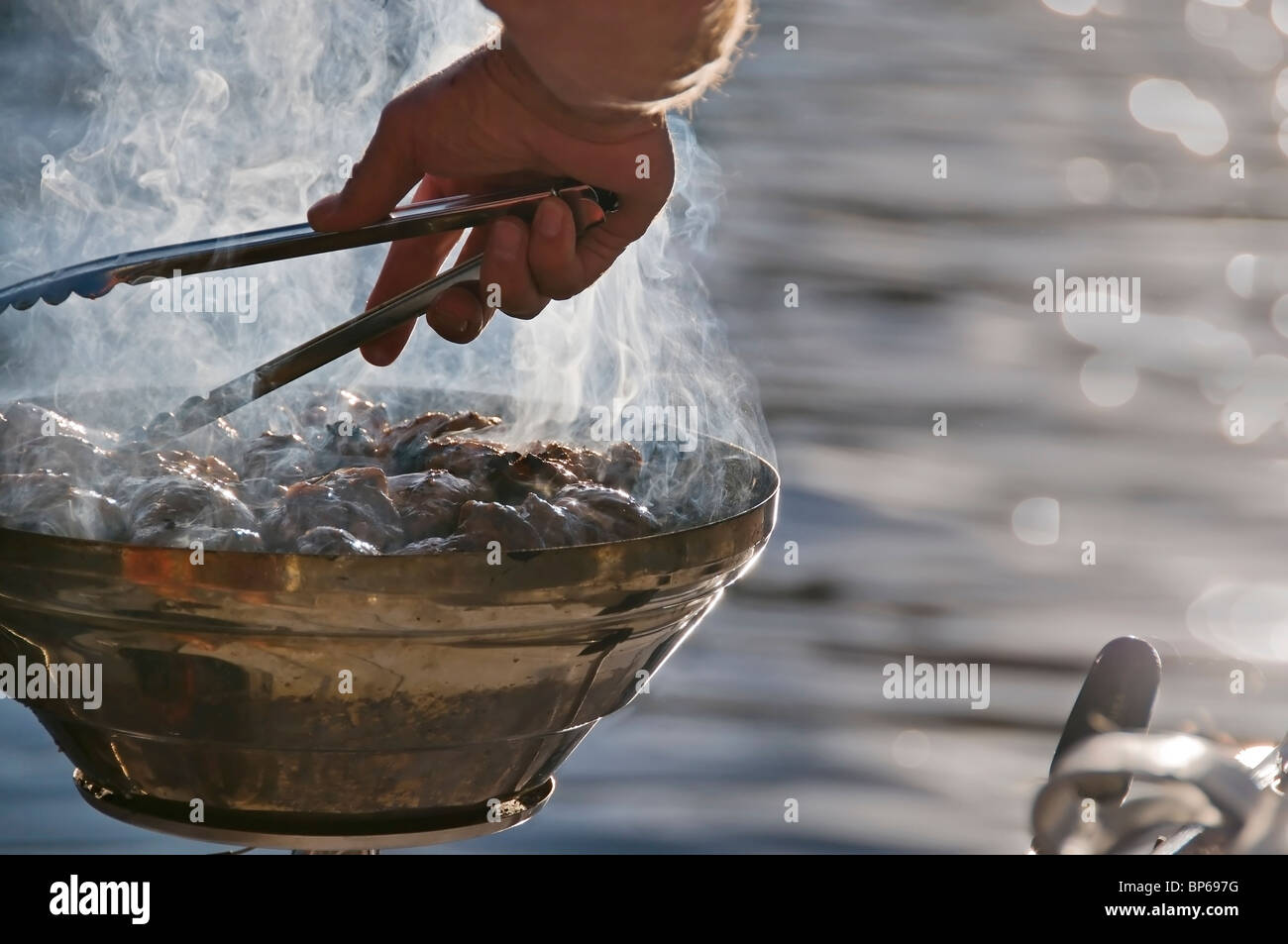 A man turns pieces of chicken with tongs over a hot barbecue grill attached to the back of a boat on the water of Puget Sound. Stock Photo