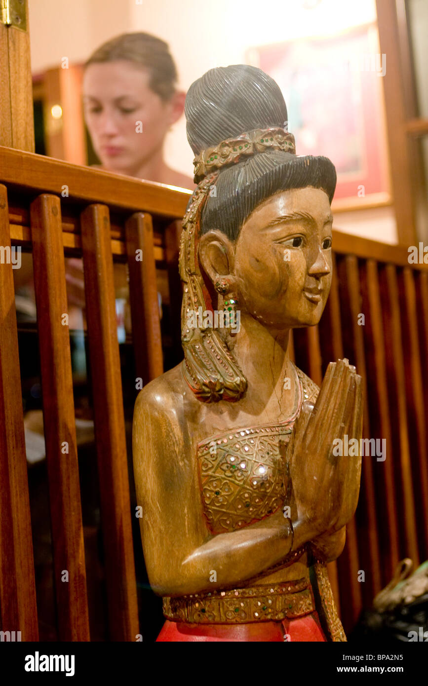 A wooden statue of a Thai woman, hands folded in a greeting wai at the