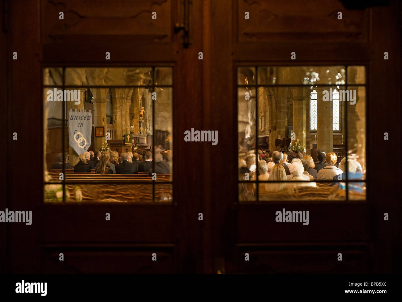 Rear view of seated congregation in UK church, seen through the windows of closed wooden doors. Stock Photo