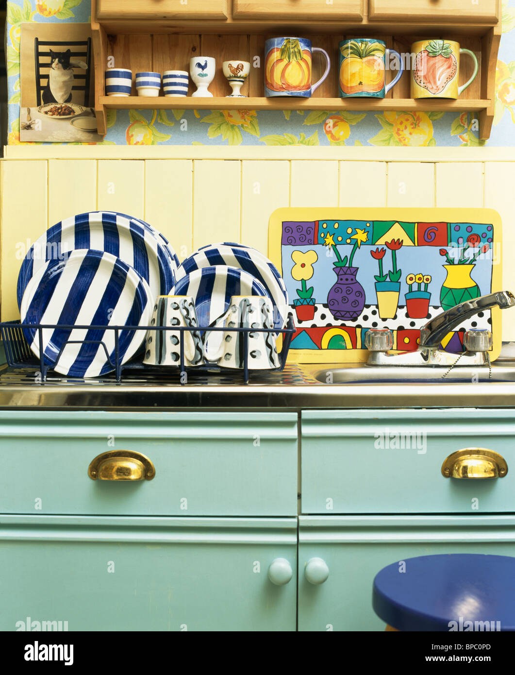 Pottery cups on pine shelf above black+white striped plates in draining rack beside sink in pale turquoise kitchen unit Stock Photo